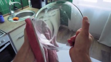 In the middle of the kitchen, near the table, a man holds a glass bowl with one hand and wipes it with a towel with the other to remove moisture. Top view from the first person. Big close up shot