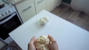 A man in the kitchen tosses a piece of dough into the air with his hands, then tears it into several pieces and throws them onto a white kitchen table. He gathers the pieces back into one piece of