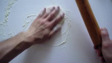 With his strong hands, the man rolls out the dough with a rolling pin on a white kitchen table sprinkled with flour. Top view from the first person on man hands. Big close up shot