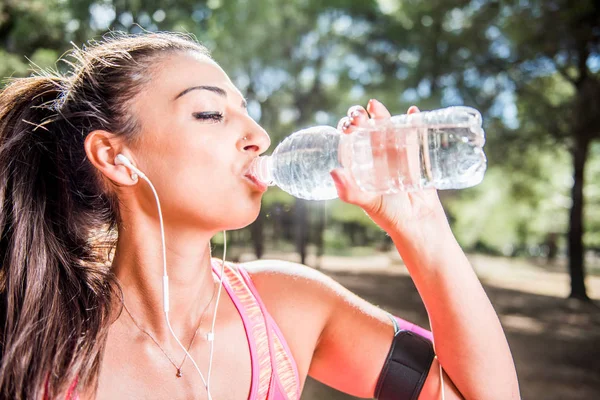 Thirsty female jogger drinking fresh water and listening music in headphones after training. Young athletic woman exercising in the park outdoors.