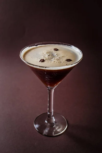 Glass of espresso martini with coffee beans and vodka on elegant dark brown background.
