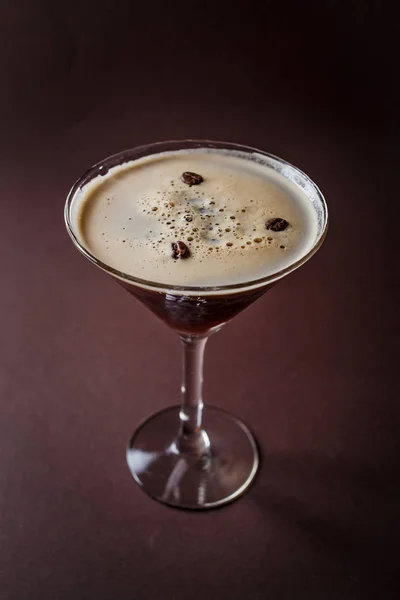 Glass of espresso martini with coffee beans and vodka on elegant dark brown background.