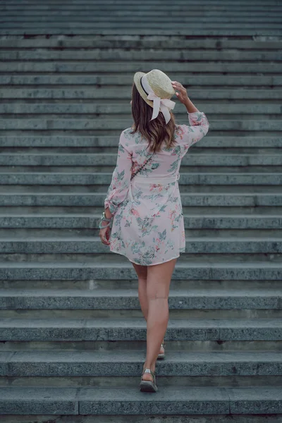 Dreaming young woman in summer dress holding hat and going up on stairways, view from her back. copy space