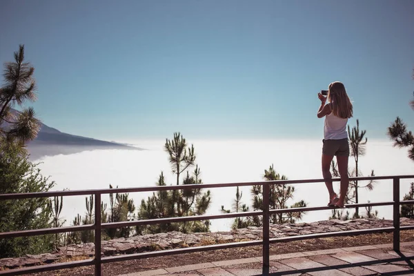 Uma jovem loira que fica no corrimão na colina sobre o céu e faz uma foto por smartphone. Sobre as montanhas e a fortaleza. Hora de verão. Estilo de vida. Vista por trás. Viajar — Fotografia de Stock