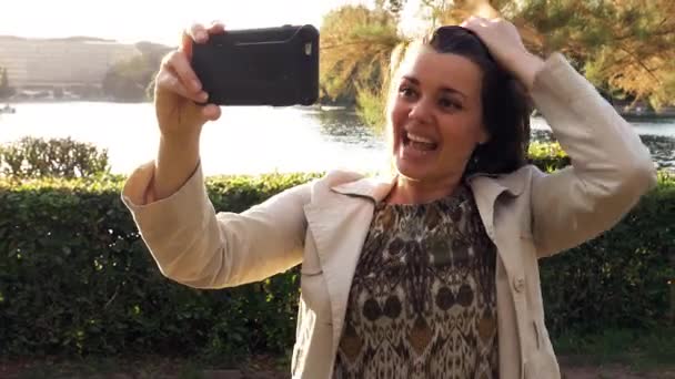 媒体を笑っているヨーロッパの都市で公園の湖からの休暇のビデオ電話を作る幸せな女はスローモーションを撮影 — ストック動画