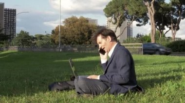 City park telefonda çimenlerin üzerinde oturan dizüstü bilgisayar ile çalışan adam