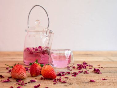 Pembe gül yaprakları, cam su ısıtıcılı, küçük, berrak bir bardakta demlenmiş çay. Ahşap kırsal arka plan.