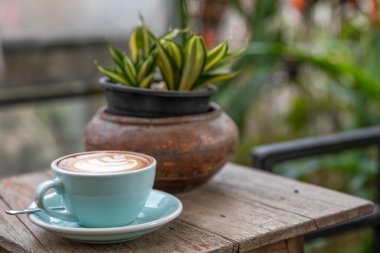 Sıcak kapuçino kahvesi, ahşap bir masada, mavi renkli seramik bardakta. Bulanık bahçe arkaplanı.