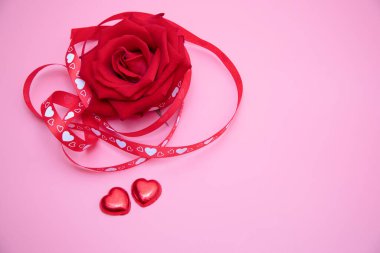 Pembe arka planda kırmızı ve beyaz kurdeleli güzel kırmızı bir gül ve iki kırmızı kalp şekeri. Sevgililer Günü, yıldönümü, düğün konsepti.