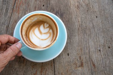 Neredeyse boş bir bardak kapuçino tutan bir adamın resmi. Gök mavisi kapsülün altında köpüklü sanat desenli. Doğal tahta masa arkaplanı.