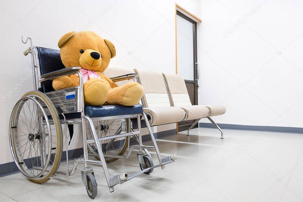 A large brown toy bear sitting on a dark blue wheel chair in a bright waiting area of a children hospital.