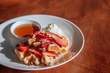 Taze çilekli çıtır waffle fırında ceviz krem şanti ve beyaz tabakta bal güneş ışığı altında ahşap masanın üstünde..
