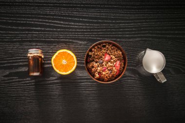 Sağlıklı kahvaltı. Tahta kasede meyveler, süt ve bal bulunan müsli siyah arka planda fotokopi alanı ile üstten görünüyor.