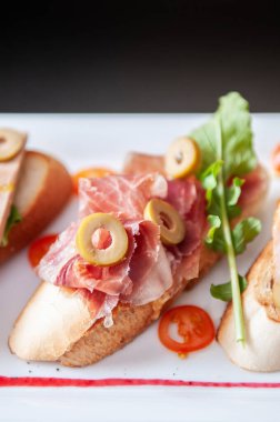 İtalyan aperatifi Carpaccio ekmekleri zeytin, Pamaham, roket marulu ve yaban mersini sosu beyaz tabakta.