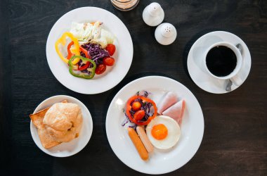 Bir set Amerikan kahvaltısı. Yumurta, sosis, jambon, taze salata dolmalığı biber, domates, sade kahveyle kruvasan ekmeği. Üst görünüm