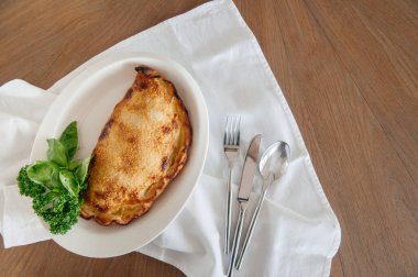 Peynirli, mantarlı, fesleğen yapraklı, maydanozlu ve ahşap masadaki gümüş takımlı Calzone pizzası. Kopyalama alanı ile üst görünüm