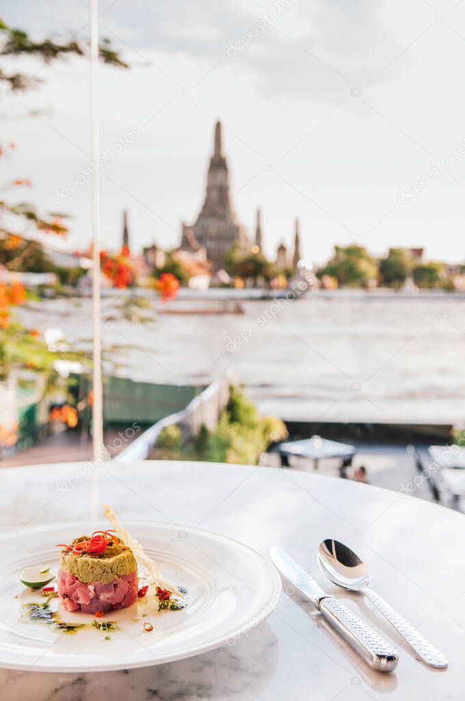 Tuna Tartare served with avocado, wasabi and lime on white plate at restaurant table with Wat Arun temple and Chao Praya river view, Bangkok, Thailand