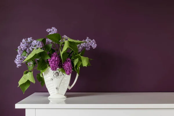 Blooming lilac flowers in the vase on purple background. Purple branches of lilac.