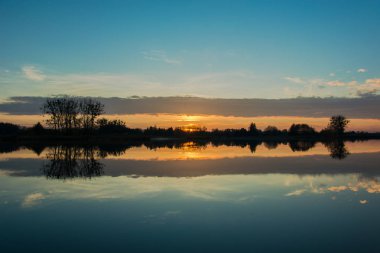 Göl, ağaçların üstünde belgili tanımlık kıyı ve yansıma bulut su üzerinde gündoğumu