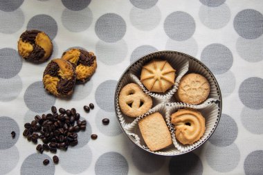 Yuvarlak bir kutu ve kahve çekirdekleri ile lekeli bir masa örtüsü üzerinde yatan üç şekerli kurabiye