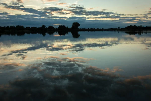 Evening clouds in the blue sky reflecting in the water of a calm lake, horizon and trees