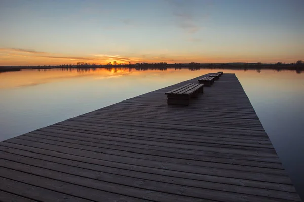 A wide wooden jetty with benches, sunset and calm lake Stock Picture