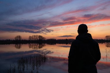 Gölün kenarındaki adam gün batımından sonra bulutlara bakıyor.
