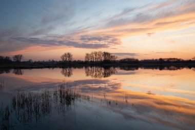Huzurlu bir göldeki renkli bulutların ve ağaçların yansıması, Mart akşamı