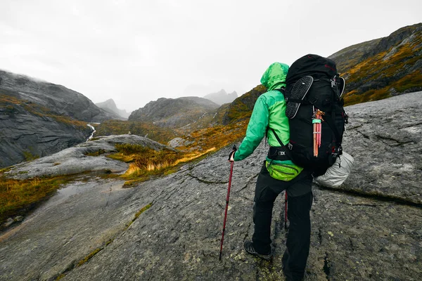 Young man hiking in the mountains of the Lofoten Islands in Norway with backpack, outdoor gear and hiking poles The hiker is taking a rest and stands on a massive rock. He is looking into the distance along the trail with his hiking poles grounded. H