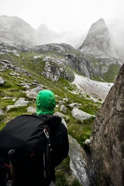 Norveç Lofoten Adası'nda dağlarda oturan bir yürüyüş adam arkasından görünümü. Dağlar kayalar ve yeşil otlarla kaplıdır. Sis ve bulutlarla yağmurlu bir gün..  