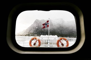 Arka planda dağlar ile Norveç Lofoten Adaları'nda bir fırtına sırasında bir feribot üzerinde norveç bayrağı ve cankurtaran bir teknenin bullseye penceresinden görünümü.