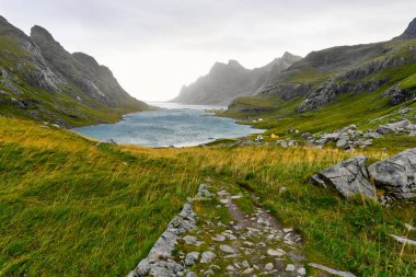 Hiking trail leading to a beautiful bay surrounded by mountains and a small village near Bunes Beach and Vinstad on Lofoten Islands in Norway clipart
