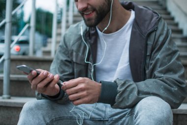 Şarkı ve şehir. Yakın çekim Genç adam otururken sanayi kenti görünümü karşı onun smartphone kullanarak Kulaklığımda parçası