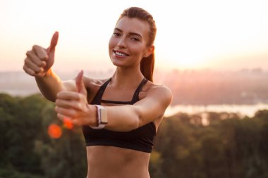 Egzersiz için en iyi yer! Genç güzel kadın gülümseyerek kameraya bakarak ve sanayi kenti görünümü karşı dururken başparmak gösterilmesini spor giyim uygun