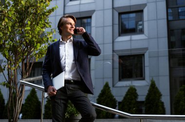 Yeni iş ortağımız 's great! Onun laptop taşıyan ve açık havada duran süre gülümseme ile onun smartphone kullanarak yakışıklı delikanlı