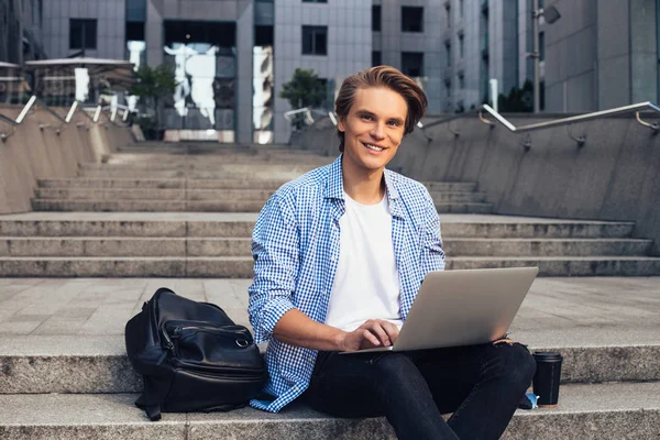 Studying can be fun! Handsome young man using his laptop and looking at camera with smile while sitting outdoors