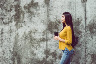 Ona kahve zevk. Uzağa bakarak ve kahve fincanı duran beton arka plan açık havada tutan güzel genç kadın yan görünüm 