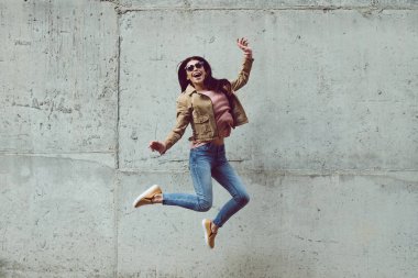 Hava tarzında!. Güzel genç kadının karşı beton arka plan açık havada atlama sırasında gülümseme ile kameraya bakarak tam uzunlukta 