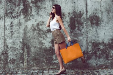 Alışveriş zamanı. Cep telefonu üzerinde konuşurken ve alışveriş torbaları duran beton arka plan açık havada tutan güzel genç kadının tam uzunlukta 