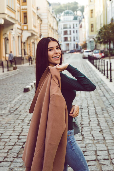 It is getting warm! Beautiful young woman in stylish coat looking at camera with smile while standing outdoors