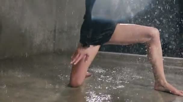 Joven bailarina en un traje negro realiza una danza contemporánea con bajo la lluvia de agua con salpicaduras en las vigas del reflector. Danza bajo la lluvia en interiores — Vídeo de stock