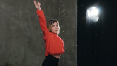 Kadın dansçı kırmızı kazaklı Studio stüdyo ışık önce su damlaları altında dans. Yağmur, seksi kız dansçı siyah şort ve kırmızı kazaklı kendini etrafında çember yapar. Dans eden kız