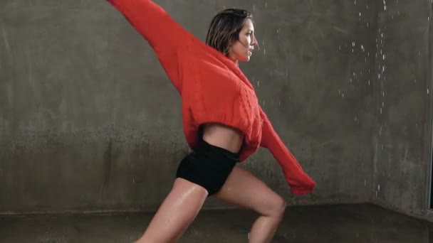Wet girl dancer in red sweater and black shorts performs contemporary dance in the rain and splashes of water. Professional dancer who danced under the water during the dance in indoors before studio — Stock Video