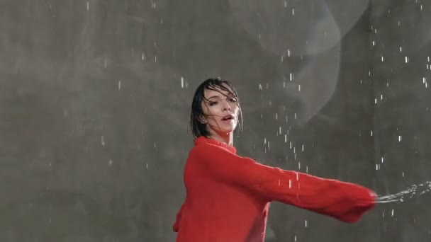 La belle blonde en pull rouge dansant sous les gouttes de pluie en studio sur fond de mur gris. La jeune danseuse exécute un élément circulaire dans la danse — Video