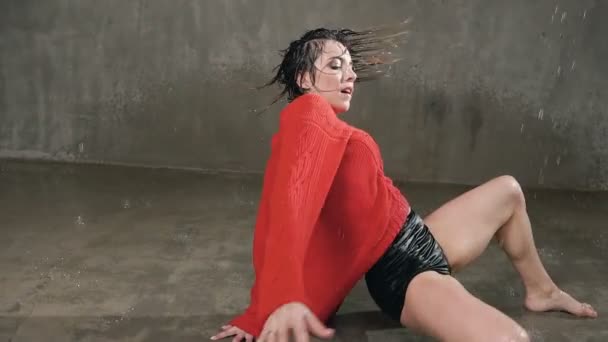 Young dancer girl in a red sweater performs a contemporary dance the floor under the water rain with splashes of water in the beams of the searchlight. Dance in the rain in indoors — Stock Video