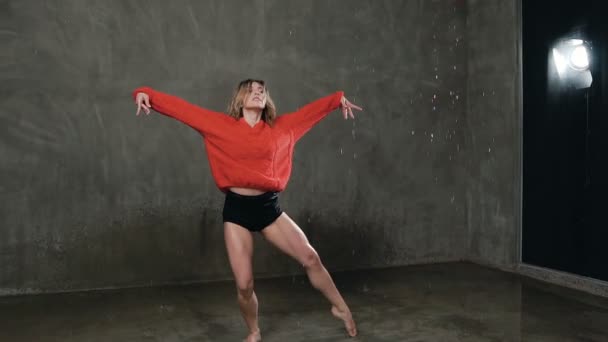 Une jeune danseuse professionnelle exécute une danse acrobatique en studio sous la pluie. Danse fille dansant la danse moderne en pull rouge et shorts sur la scène en gouttes d'eau — Video