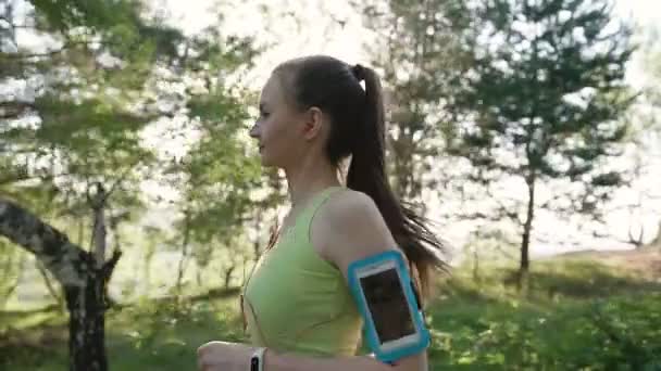 Atletica giovane donna caucasica che corre e ascolta musica in cuffia su smartphone all'aperto nella foresta al tramonto. Atletica ragazza fa una corsetta nel parco mentre ascolta la musica — Video Stock