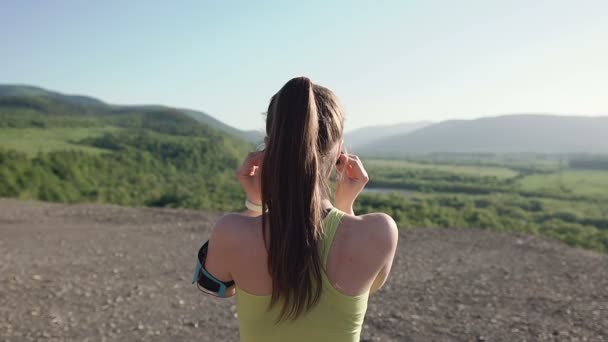 Vue arrière. Fille athlète jogging dans les montagnes au coucher du soleil en cours d'exécution de la caméra et écouter de la musique dans le casque sur un smartphone. Femme sportive écoutant de la musique pendant l'entraînement de course en soirée — Video