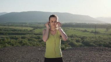 Kamera, bir koşu dağlarda günbatımı, o giyer telefondan müzik dinlemek için kulaklık yapar bir kız atlet binmek. Çekici spor kız lisening müzik için kulaklık takıyor