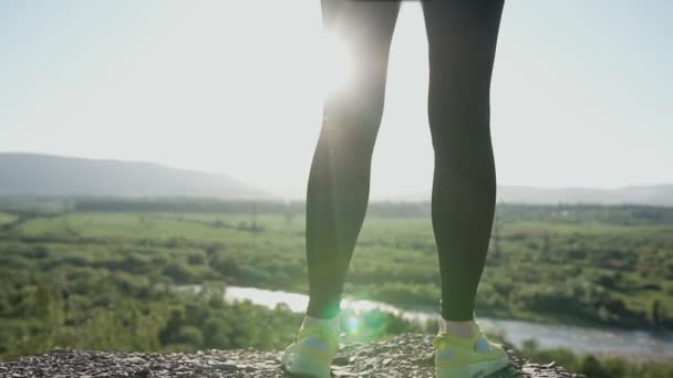 Chiudete. Giovane donna sportiva in piedi sul bordo della scogliera e brillante foresta verde estiva al tramonto. Atletica ragazza sportiva godendo ascoltare musica ha smartphone e cuffie — Video Stock